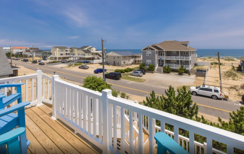 View from a balcony overlooking a beachside street with houses and the ocean in the distance under a clear blue sky.