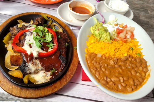A sizzling fajita platter with grilled meat, peppers, and onions, served with rice, beans, guacamole, and salsa.