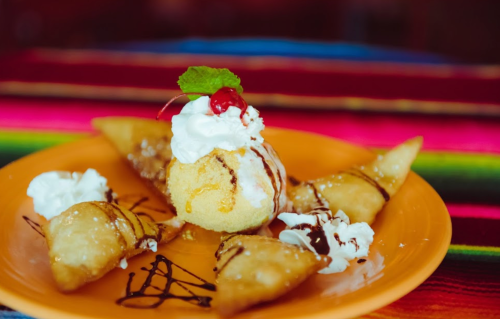 A vibrant plate of fried pastries with ice cream, whipped cream, a cherry, and chocolate drizzle on a colorful tablecloth.