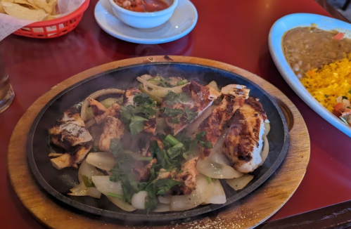 Sizzling chicken fajitas with onions and cilantro on a hot skillet, served with salsa and sides in a restaurant setting.