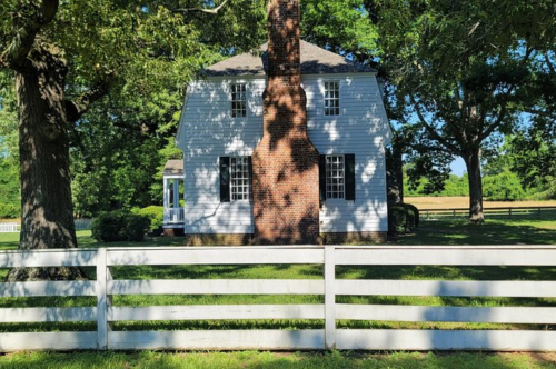 A white house with a brick chimney, surrounded by trees and a white fence, set in a green landscape.