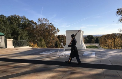 A soldier in uniform walks past a memorial, holding a rifle, with trees and a clear sky in the background.