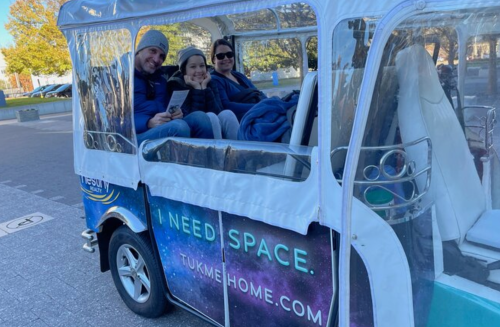 Three people sit in a covered tuk-tuk, smiling and bundled up, with a colorful "I Need Space" sign on the side.