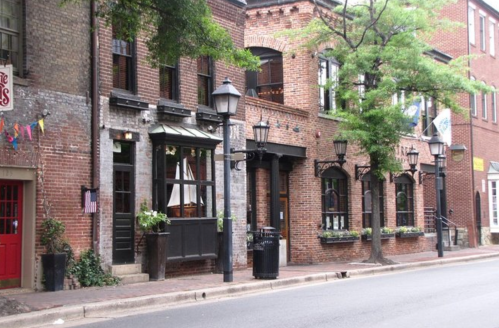 A charming street view of brick buildings with outdoor seating and lampposts, lined with trees.