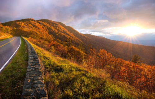A scenic view of a winding road beside vibrant autumn foliage and mountains, with a sunset illuminating the sky.