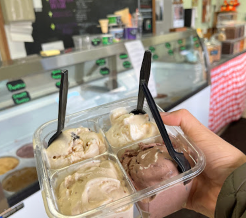 A hand holding a container with four scoops of ice cream, with a shop display in the background.
