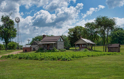 A rustic log cabin and outbuildings surrounded by green grass and trees, with a windmill in the background under a cloudy sky.