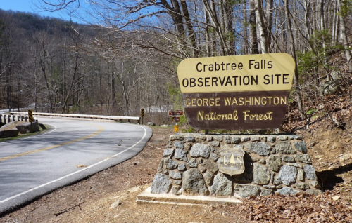 Sign for Crabtree Falls Observation Site in George Washington National Forest, with a winding road in the background.