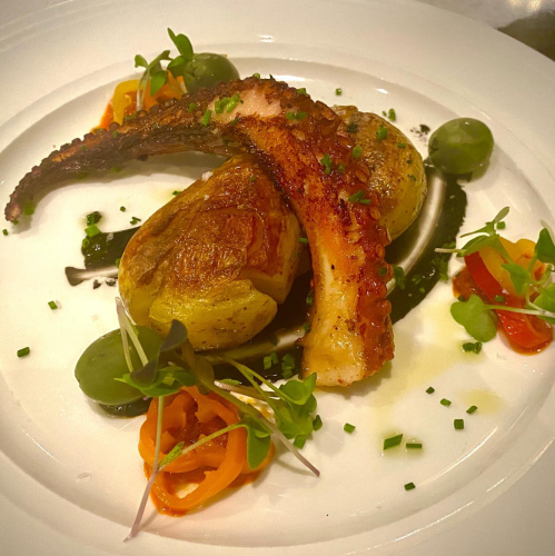 A plated dish featuring grilled octopus, roasted potatoes, and colorful vegetable garnishes on a white plate.