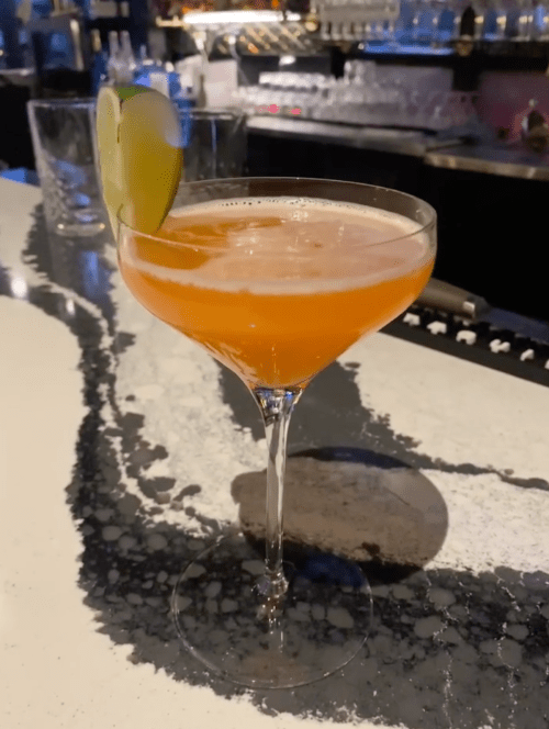 A cocktail in a coupe glass, garnished with a lime wedge, sits on a marble bar top.