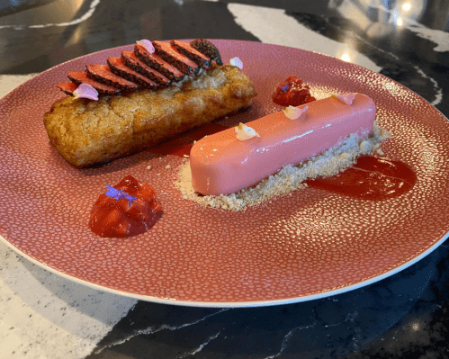 A pink dessert and a slice of grilled meat on a decorative plate, garnished with sauce and edible flowers.