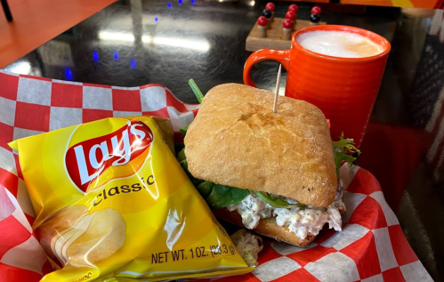 A sandwich with chicken salad and greens, a bag of Lay's chips, and a cup of coffee on a checkered tablecloth.