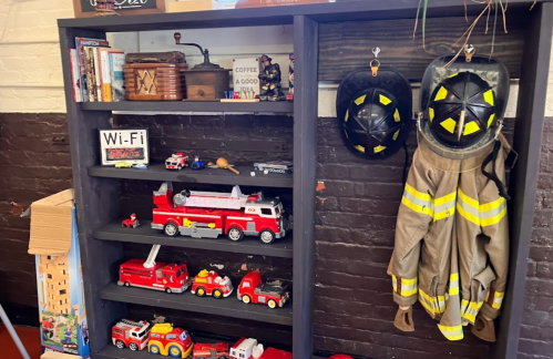 A shelf with toy fire trucks, books, and a firefighter's coat and helmet hanging on the wall.