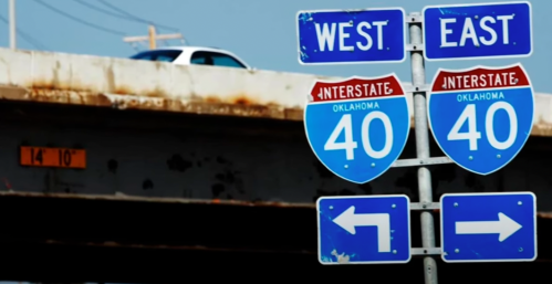 Interstate 40 signs indicating directions for West and East in Oklahoma, with a highway overpass in the background.