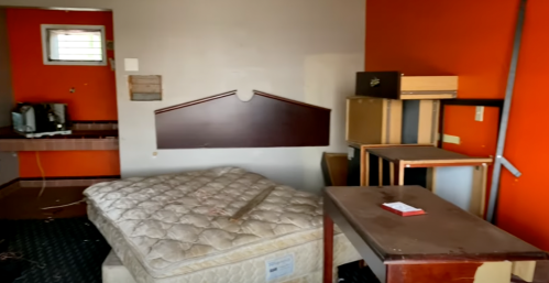 A cluttered room with an unmade bed, scattered furniture, and orange walls, suggesting neglect or abandonment.
