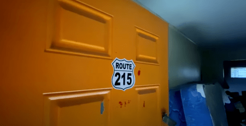 An orange door with a "Route 215" sign, showing signs of wear and some red stains, in a dimly lit room.