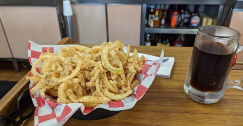 A basket of crispy onion rings on a checkered paper, next to a glass of dark soda on a wooden table.