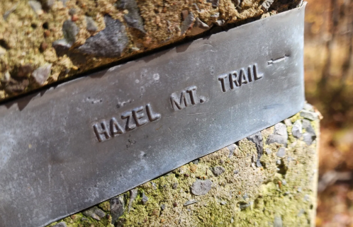 Metal sign reading "HAZEL MT. TRAIL" affixed to a moss-covered stone surface.