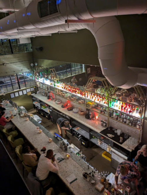 Aerial view of a stylish bar with colorful bottles, patrons seated, and a bartender preparing drinks.