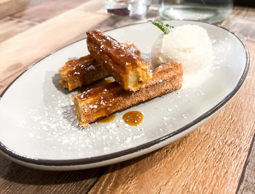 Three churros drizzled with caramel sauce, served with a scoop of ice cream on a white plate.
