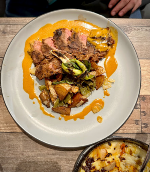 A plate of sliced beef with a sauce, roasted potatoes, and grilled vegetables on a wooden table.