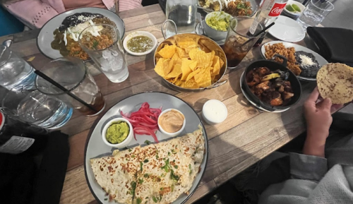 A table spread with various dishes, including nachos, tacos, and sauces, alongside drinks in glasses.