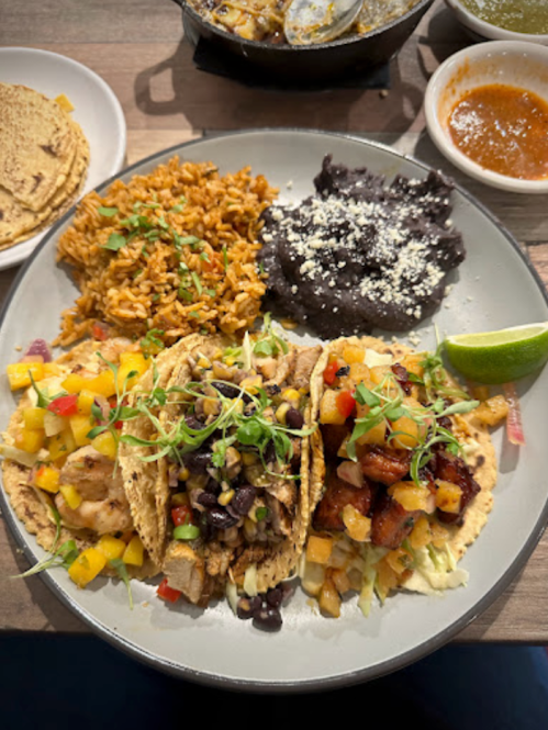 A plate of three tacos topped with various ingredients, served with rice, black beans, and a lime wedge.
