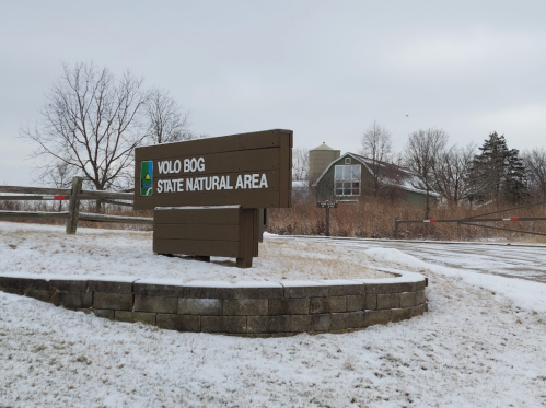 Sign for Volo Bog State Natural Area, with a snowy landscape and a building in the background.