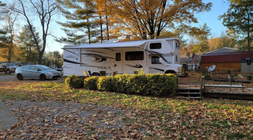 A white RV parked near a wooden deck, surrounded by autumn leaves and trees with colorful foliage.