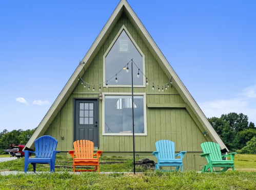 A green A-frame house with colorful chairs in front, set against a clear blue sky and grassy area.