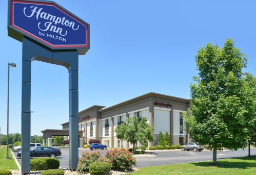A Hampton Inn by Hilton sign in front of a modern hotel building surrounded by green trees and clear blue sky.