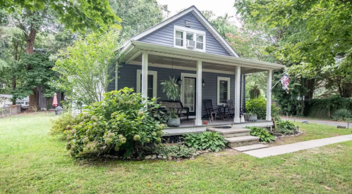 A charming blue house with a front porch, surrounded by greenery and flowers in a peaceful setting.