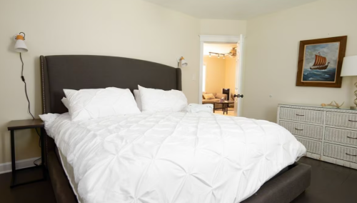 A cozy bedroom featuring a large bed with white bedding, a nightstand, and a dresser, with a doorway to another room.