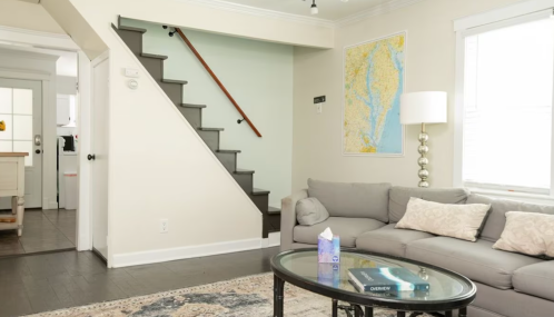 A cozy living room with a gray sofa, a glass coffee table, and a map on the wall, featuring stairs in the background.