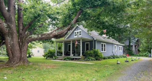 A charming blue house with a porch, surrounded by greenery and large trees, set in a peaceful rural landscape.