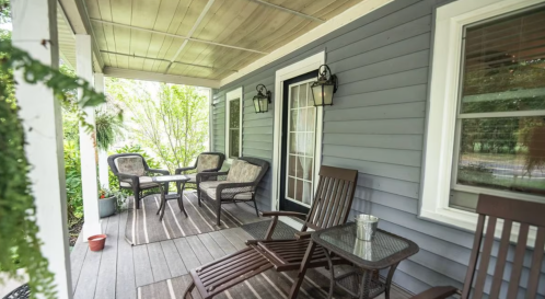 A cozy porch with seating, including chairs and a table, surrounded by greenery and natural light.