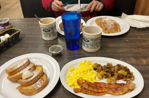 A breakfast spread featuring French toast, scrambled eggs, bacon, hash browns, coffee, and pancakes on a wooden table.