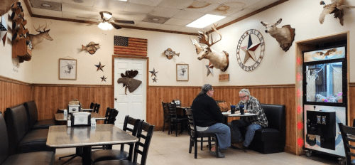 Two people sit at a table in a rustic diner decorated with mounted animal heads and wooden paneling.