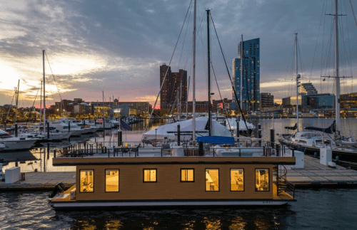 A modern houseboat docked at a marina during sunset, surrounded by yachts and city buildings.