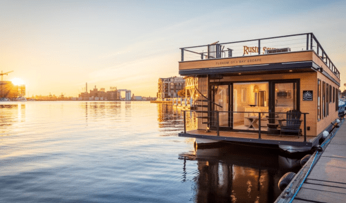 A modern houseboat at sunset, reflecting on calm waters, with city buildings in the background.