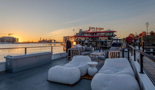A cozy outdoor seating area on a boat, overlooking a sunset and the Rusty Sipper restaurant in the background.