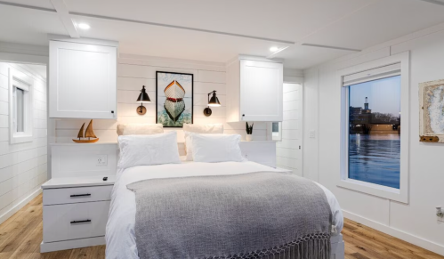 Cozy bedroom with a bed, white walls, and large windows overlooking water, featuring nautical decor and warm lighting.