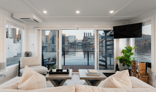 Cozy living area with large windows overlooking a waterfront view, featuring modern furniture and plants.