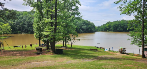 A serene lake surrounded by trees, with people walking along the shore and benches in a grassy area.