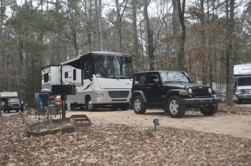 A black Jeep parked next to a large RV in a wooded campsite with fallen leaves on the ground.