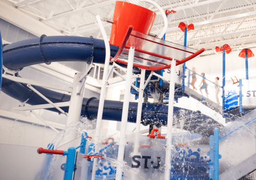 A colorful indoor water slide with a large bucket pouring water, surrounded by playful water features and children.
