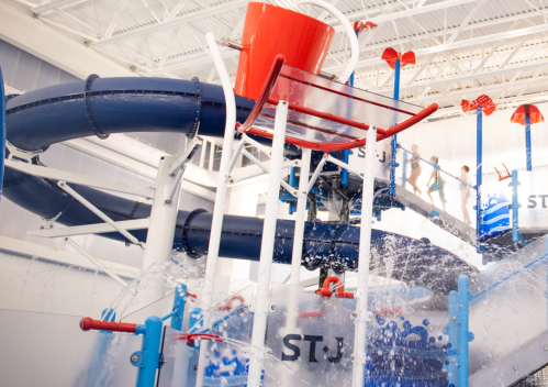 A colorful indoor water slide with a large bucket pouring water, surrounded by playful water features and children.