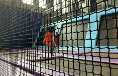 Two people jumping on trampolines, viewed through a safety net in a trampoline park.