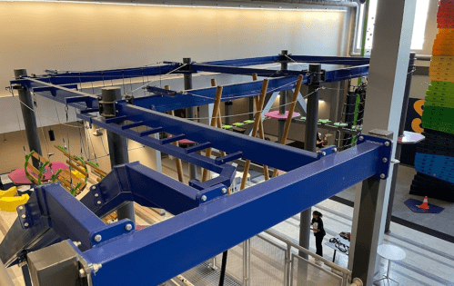 A colorful indoor climbing structure with ropes and platforms, surrounded by a spacious area and people below.