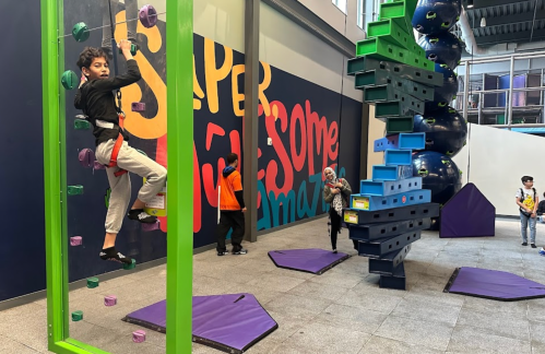 A child climbs a colorful rock wall while others play nearby in a vibrant indoor activity space.